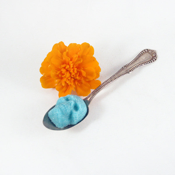 Turquoise Sugar Cube Skull, on silver spoon, next to orange marigold, white background.