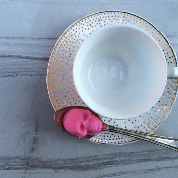 DemBones, hot pink sugar cube skull, on spoon, white and gold tea cup, marble table.