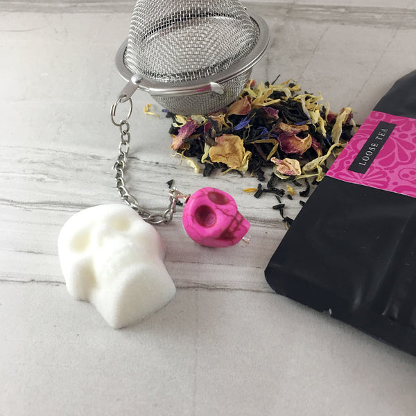 Dried Rose Marigold and Cornflower Tea spilling out of tea ball on light tile, Skull sugar cubes , Black and pink bag with words Loose Tea